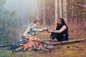 Two women sharing tea and warmth by a bonfire in a serene forest setting.