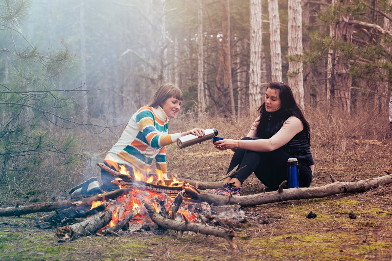 Two women sharing tea and warmth by a bonfire in a serene forest setting.